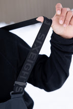 A person in a black long-sleeve shirt holds a black strap with "SIX EIGHT S" from the 686 Men's GORE-TEX Dispatch™ Shell Bib. The snowy background is blurred.