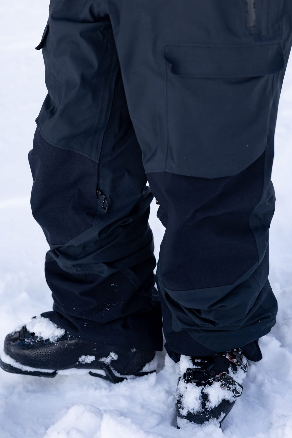A person wearing the 686 Men's GORE-TEX Dispatch™ Shell Bib stands on snow, with flakes clinging to the black bib and boots.
