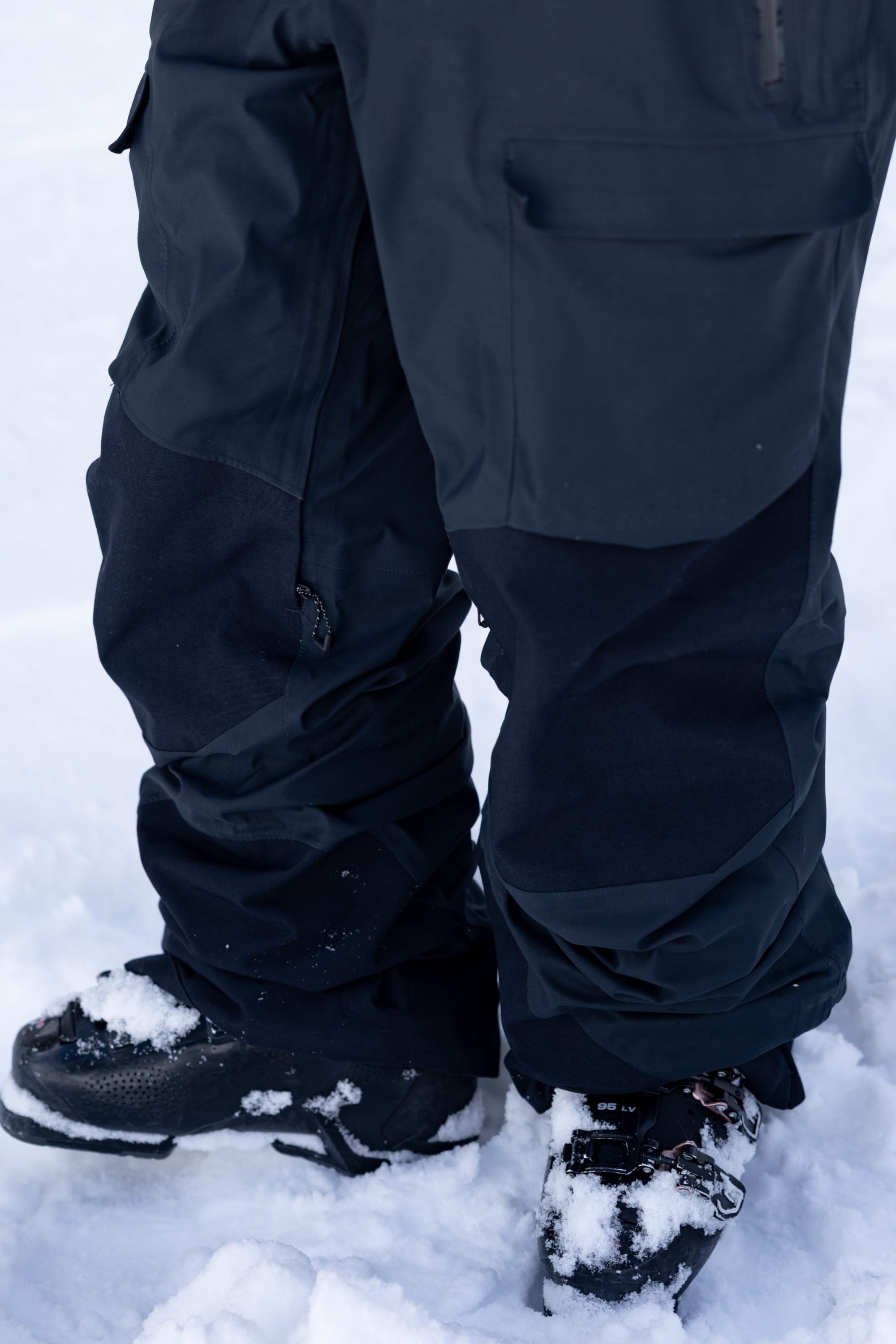 A person wearing the 686 Men's GORE-TEX Dispatch™ Shell Bib stands on snow, with flakes clinging to the black bib and boots.