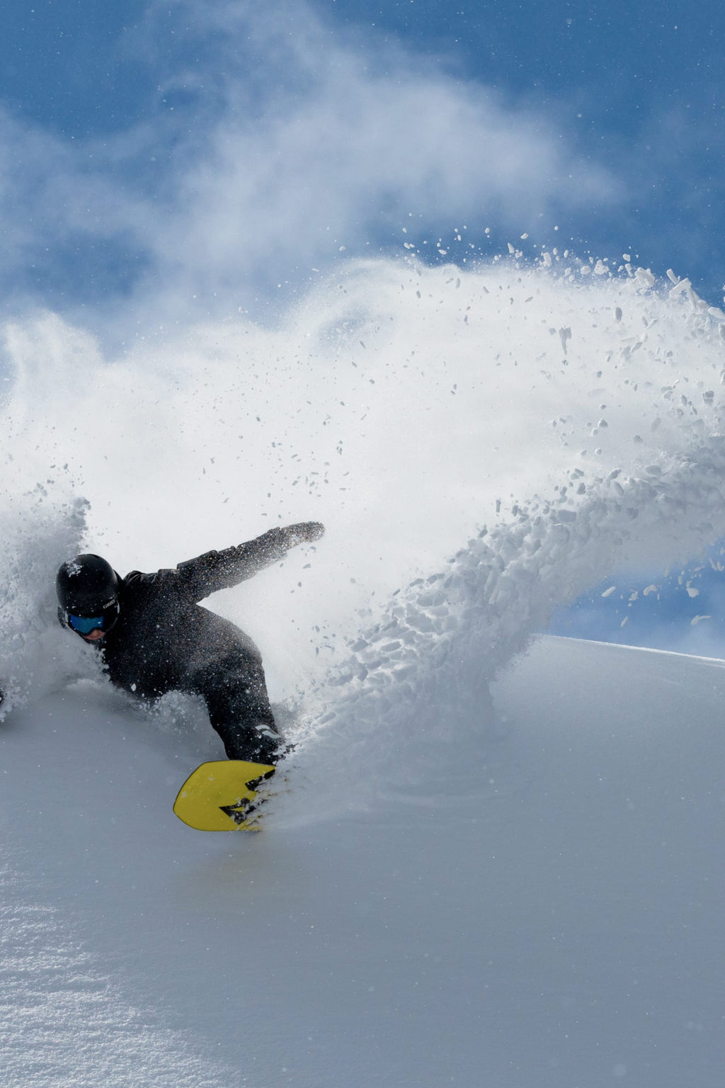 A snowboarder wearing the 686 Men's GORE-TEX Dispatch™ Shell Bib by 686 and a helmet carves through fresh snow, kicking up a dramatic spray of powder beneath a clear blue sky.