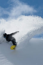 A snowboarder wearing the 686 Men's GORE-TEX Dispatch™ Shell Bib by 686 and a helmet carves through fresh snow, kicking up a dramatic spray of powder beneath a clear blue sky.