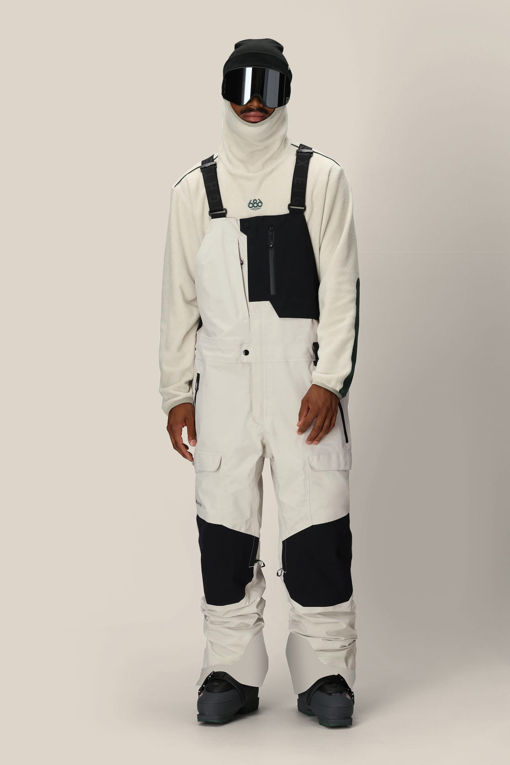 Wearing the 686 Men's GORE-TEX Dispatch™ Shell Bib in black and white with a white fleece pullover, black snow boots, gloves, ski goggles, and a balaclava, a person stands against a plain light background.