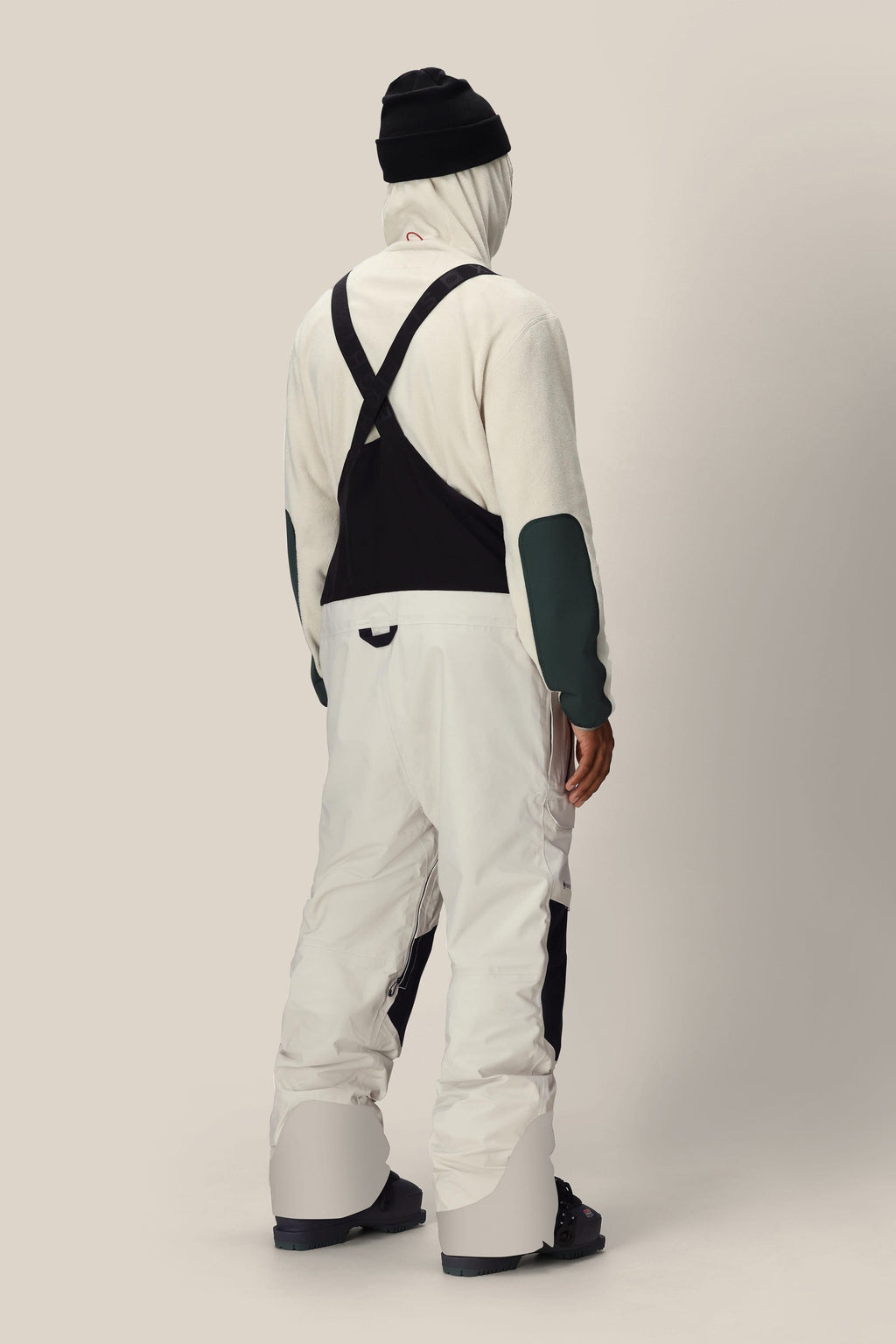 A person stands facing away in white 686 Men's GORE-TEX Dispatch™ Shell Bibs with black suspenders, black boots, and a black beanie against a plain light background.