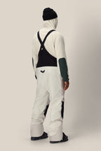 A person stands facing away in white 686 Men's GORE-TEX Dispatch™ Shell Bibs with black suspenders, black boots, and a black beanie against a plain light background.
