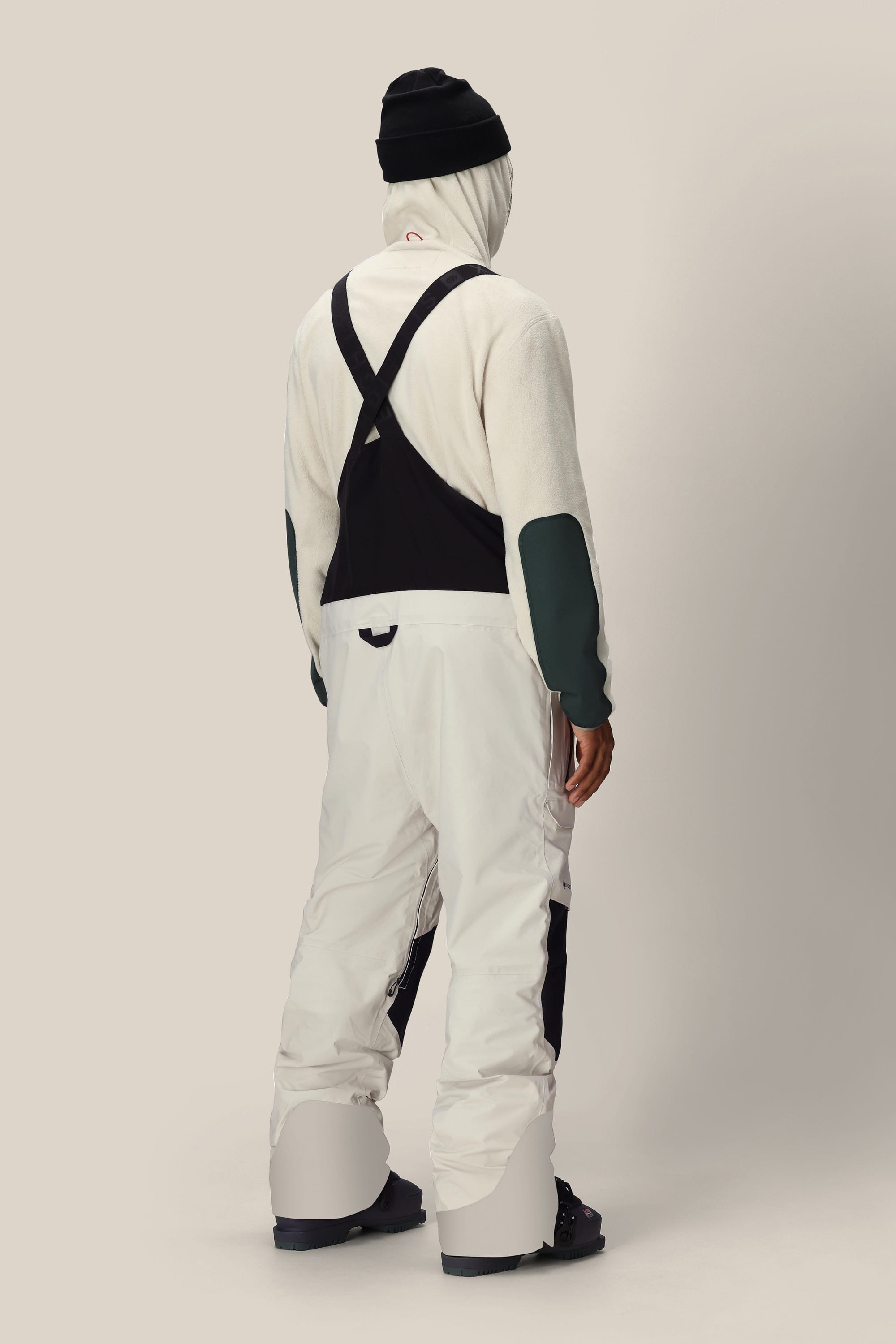 A person stands facing away in white 686 Men's GORE-TEX Dispatch™ Shell Bibs with black suspenders, black boots, and a black beanie against a plain light background.