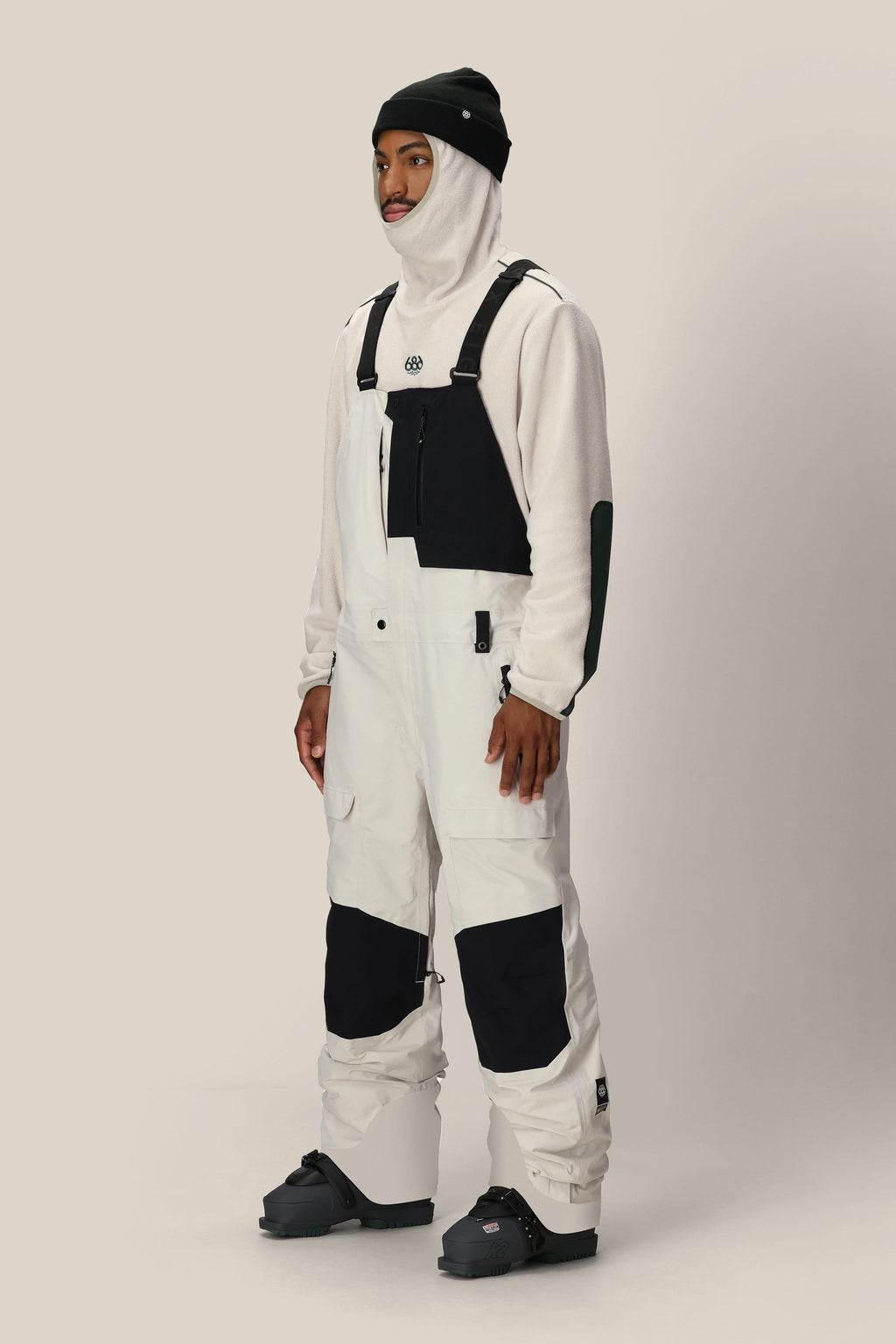 A person wearing a beanie, balaclava, snow boots, and the 686 Men's GORE-TEX Dispatch™ Shell Bib from 686 stands against a plain light background.