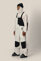 A person wearing a beanie, balaclava, snow boots, and the 686 Men's GORE-TEX Dispatch™ Shell Bib from 686 stands against a plain light background.