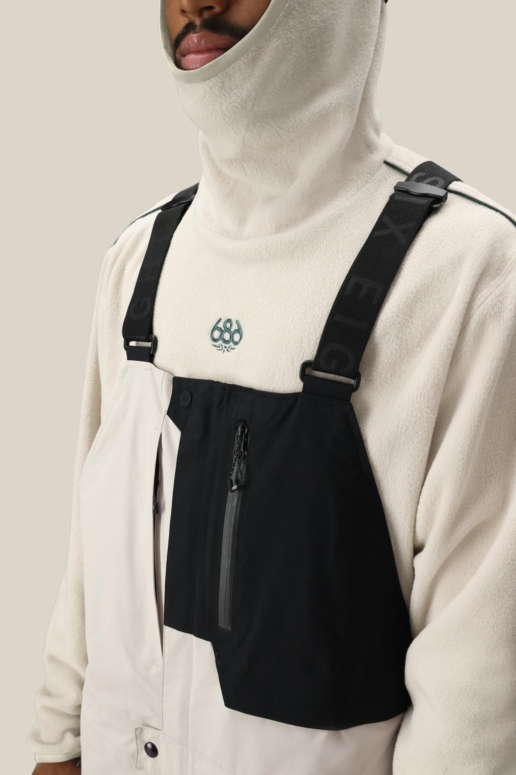 Wearing a cream fleece hooded top with a black chest panel and front zipper pocket, secured by black text suspenders, styled over the 686 Men's GORE-TEX Dispatch™ Shell Bib. Only the lower half of the face is visible.