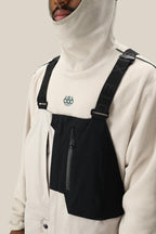Wearing a cream fleece hooded top with a black chest panel and front zipper pocket, secured by black text suspenders, styled over the 686 Men's GORE-TEX Dispatch™ Shell Bib. Only the lower half of the face is visible.