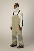 A person wears the 686 Men's GORE-TEX Dispatch™ Shell Bib by 686 in beige, styled with a white fleece featuring dark elbow patches, a white balaclava, and a striped beanie, standing against a plain light background.