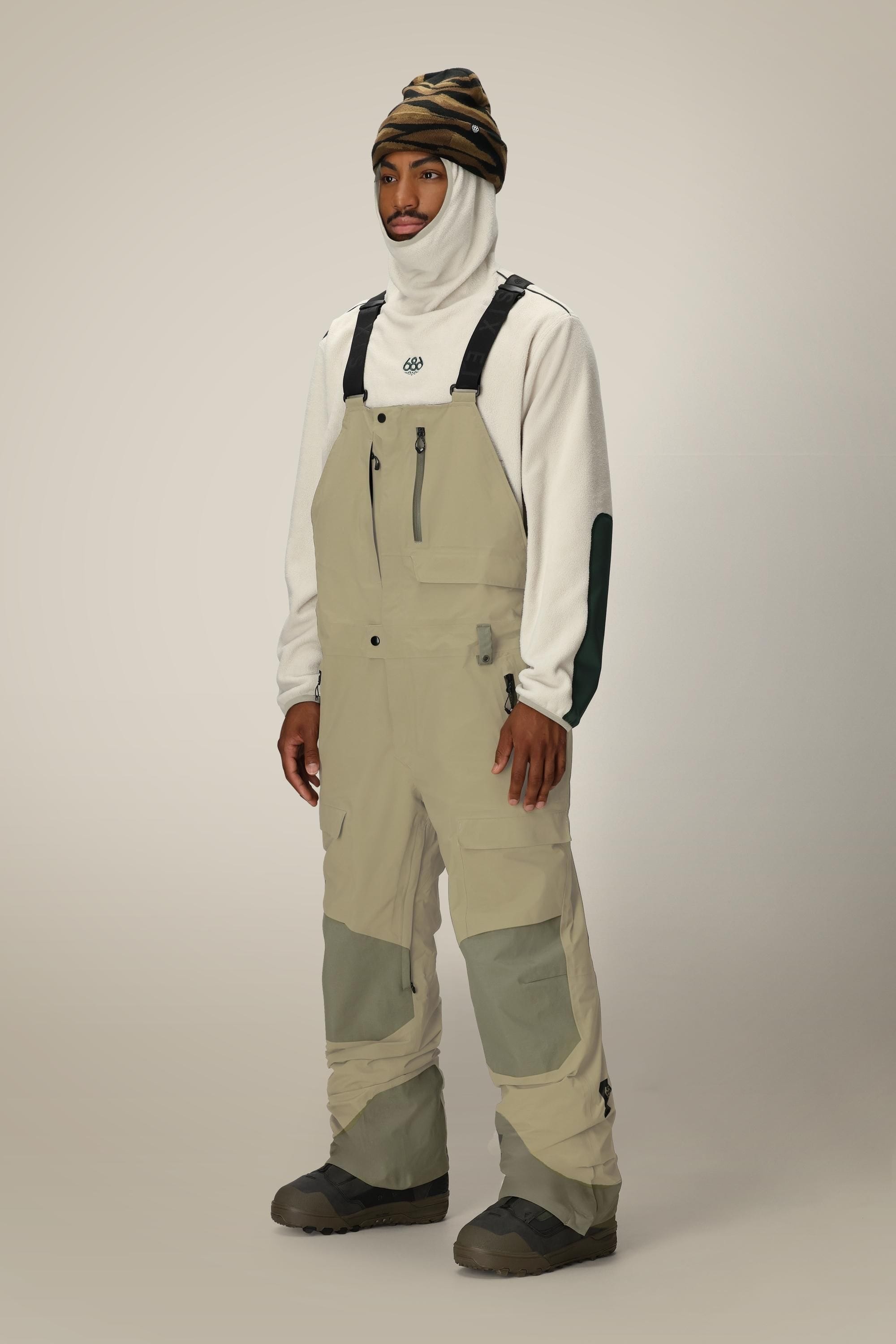 A person wears the 686 Men's GORE-TEX Dispatch™ Shell Bib by 686 in beige, styled with a white fleece featuring dark elbow patches, a white balaclava, and a striped beanie, standing against a plain light background.