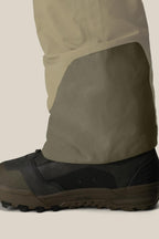 A close-up of a person's lower leg in tan pants with reinforced fabric over brown and black outdoor shoes, paired with the 686 Men's GORE-TEX Dispatch™ Shell Bib by 686, set against a light background.
