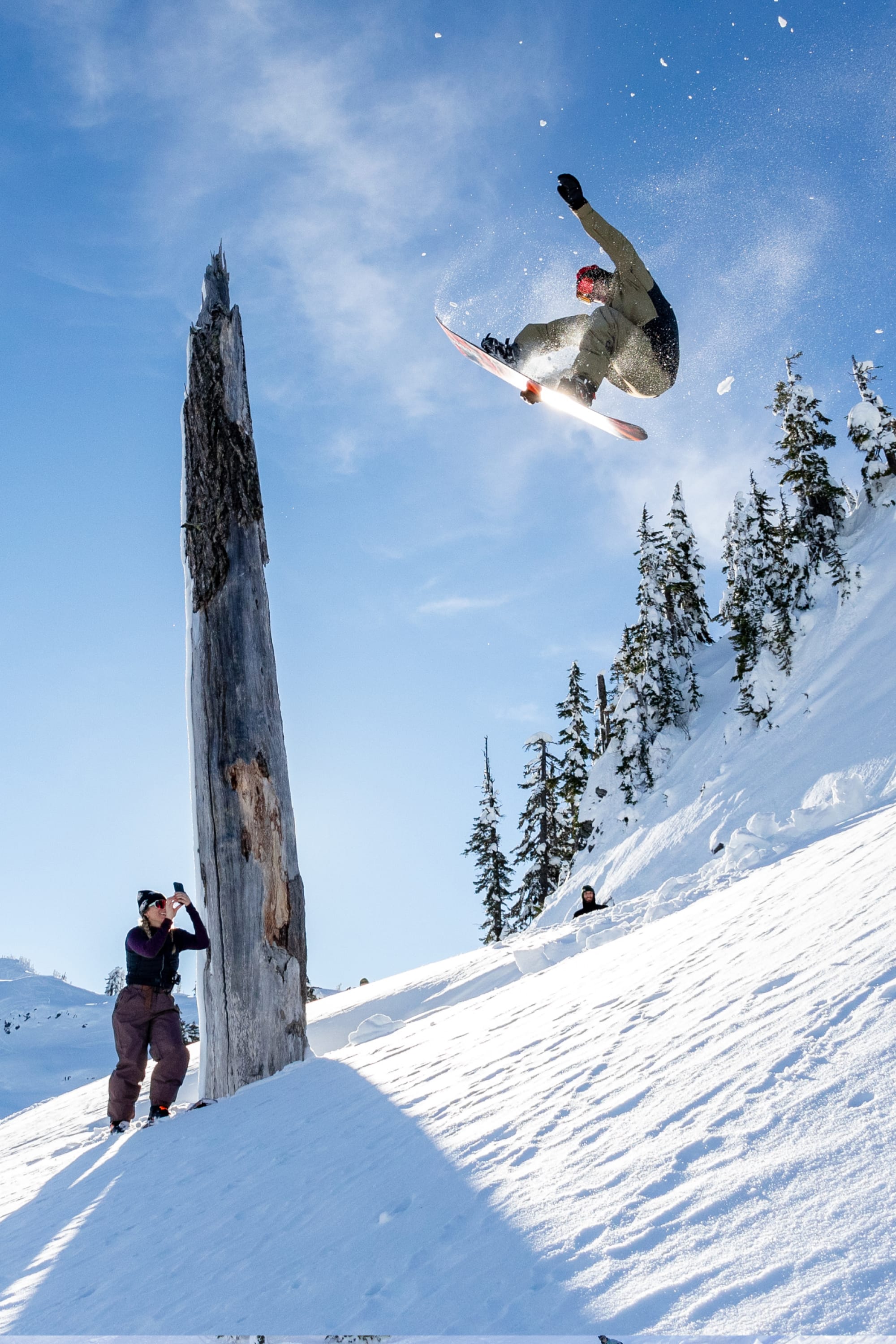 A snowboarder soars above a snowy slope as someone in the 686 Men's GORE-TEX Dispatch™ Shell Bib captures the moment; snow-covered trees and a clear blue sky fill the background.