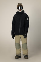 A person stands against a plain light background wearing the 686 Men's GORE-TEX Dispatch™ Shell Bib from 686, paired with a brown striped beanie, large ski goggles, black jacket, black gloves, and snow boots.