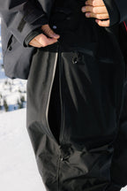 Wearing the 686 Mens GORE-TEX 3L ATV Bib, a person stands outdoors in the snow and unzips a large side pocket or vent on their black snow pants.
