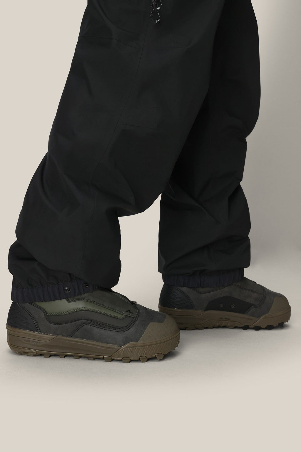Someone stands on a light surface wearing 686 Mens GORE-TEX 3L ATV Bib pants by 686, their loose black legs draping over dark green and black sneakers, slightly covering the tops.