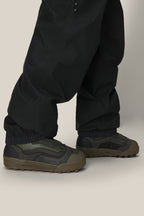 Someone stands on a light surface wearing 686 Mens GORE-TEX 3L ATV Bib pants by 686, their loose black legs draping over dark green and black sneakers, slightly covering the tops.
