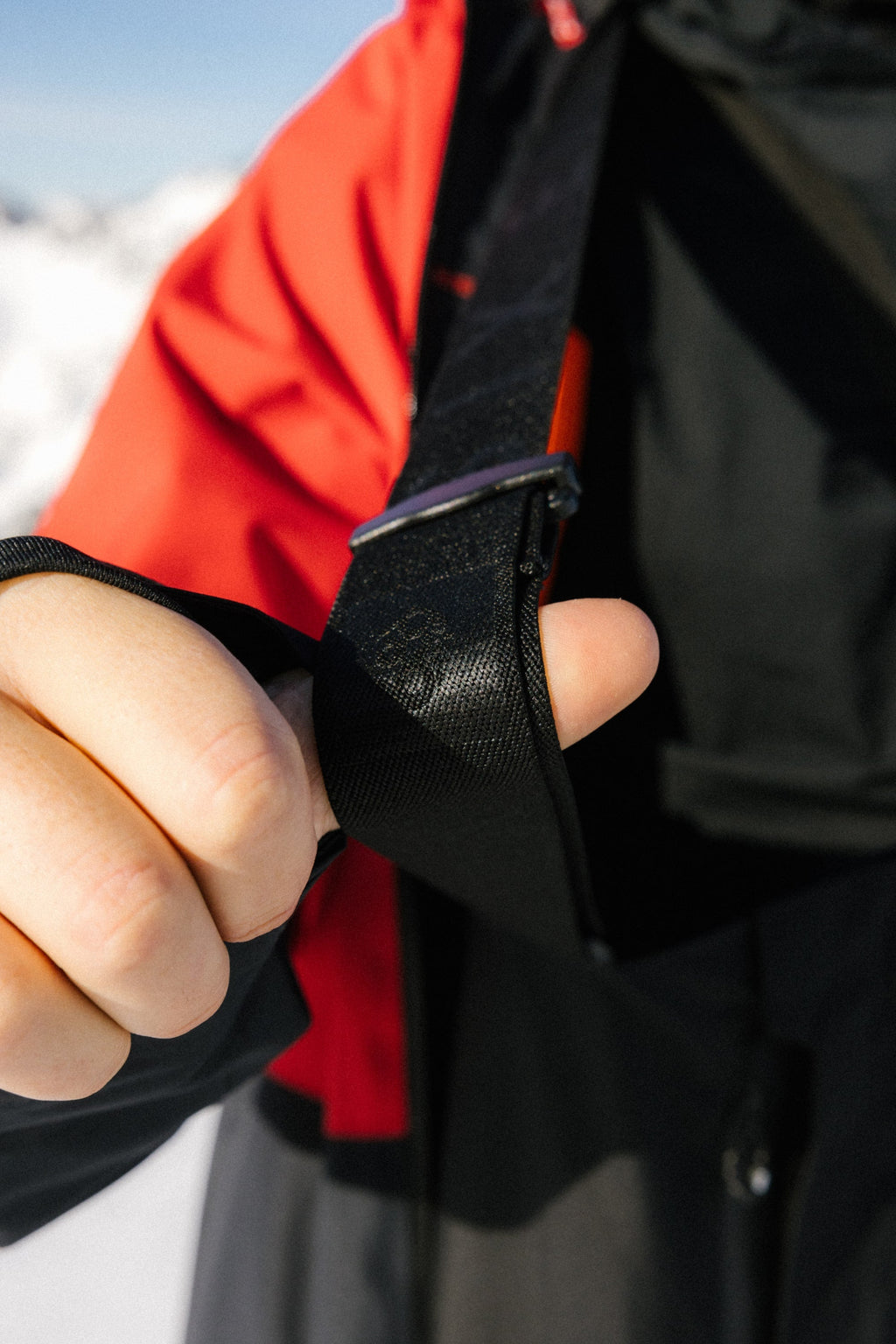 A close-up of a hand adjusting the black strap on a 686 Mens GORE-TEX 3L ATV Bib, with its red and black sleeve visible. Snowy mountains and a blue sky blur in the background.