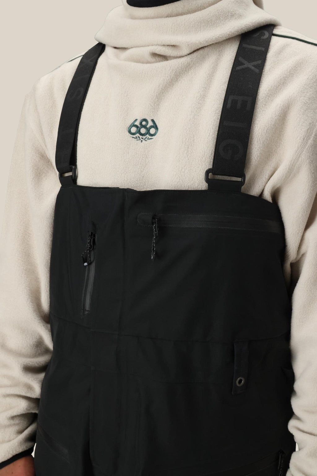 A person wears a light beige fleece pullover with an embroidered chest graphic under 686 Mens GORE-TEX 3L ATV Bib overalls, featuring zippered pockets and SIX EIGHT branding on the straps.