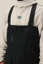 A person wears a light beige fleece pullover with an embroidered chest graphic under 686 Mens GORE-TEX 3L ATV Bib overalls, featuring zippered pockets and SIX EIGHT branding on the straps.
