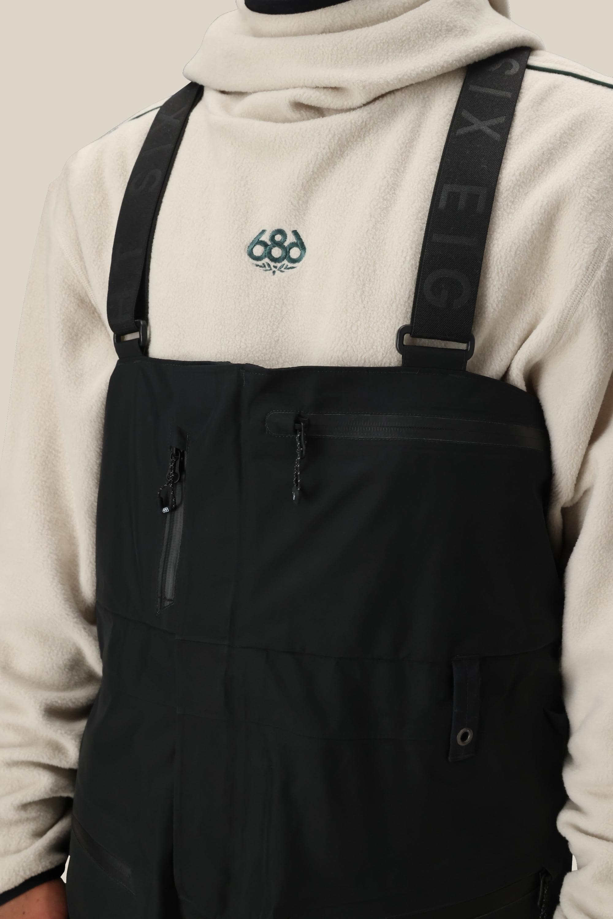 A person wears a light beige fleece pullover with an embroidered chest graphic under 686 Mens GORE-TEX 3L ATV Bib overalls, featuring zippered pockets and SIX EIGHT branding on the straps.
