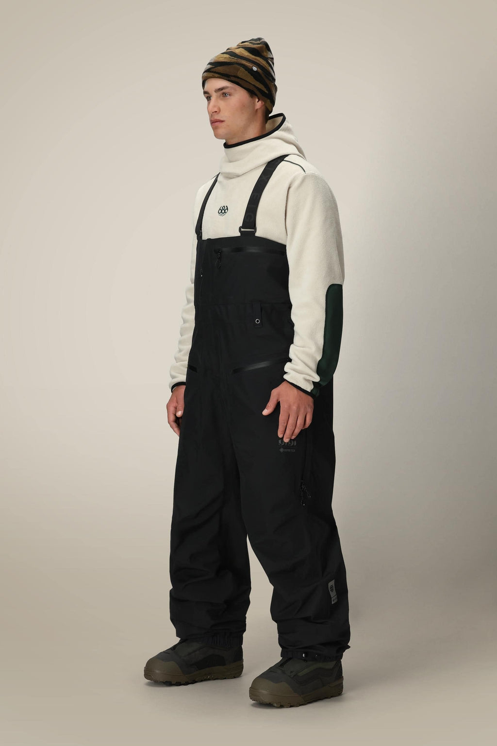 A person stands against a plain beige background wearing a white hoodie with black patches on the sleeves, 686 Mens GORE-TEX 3L ATV Bib snow overalls by 686, brown winter boots, and a striped beanie.