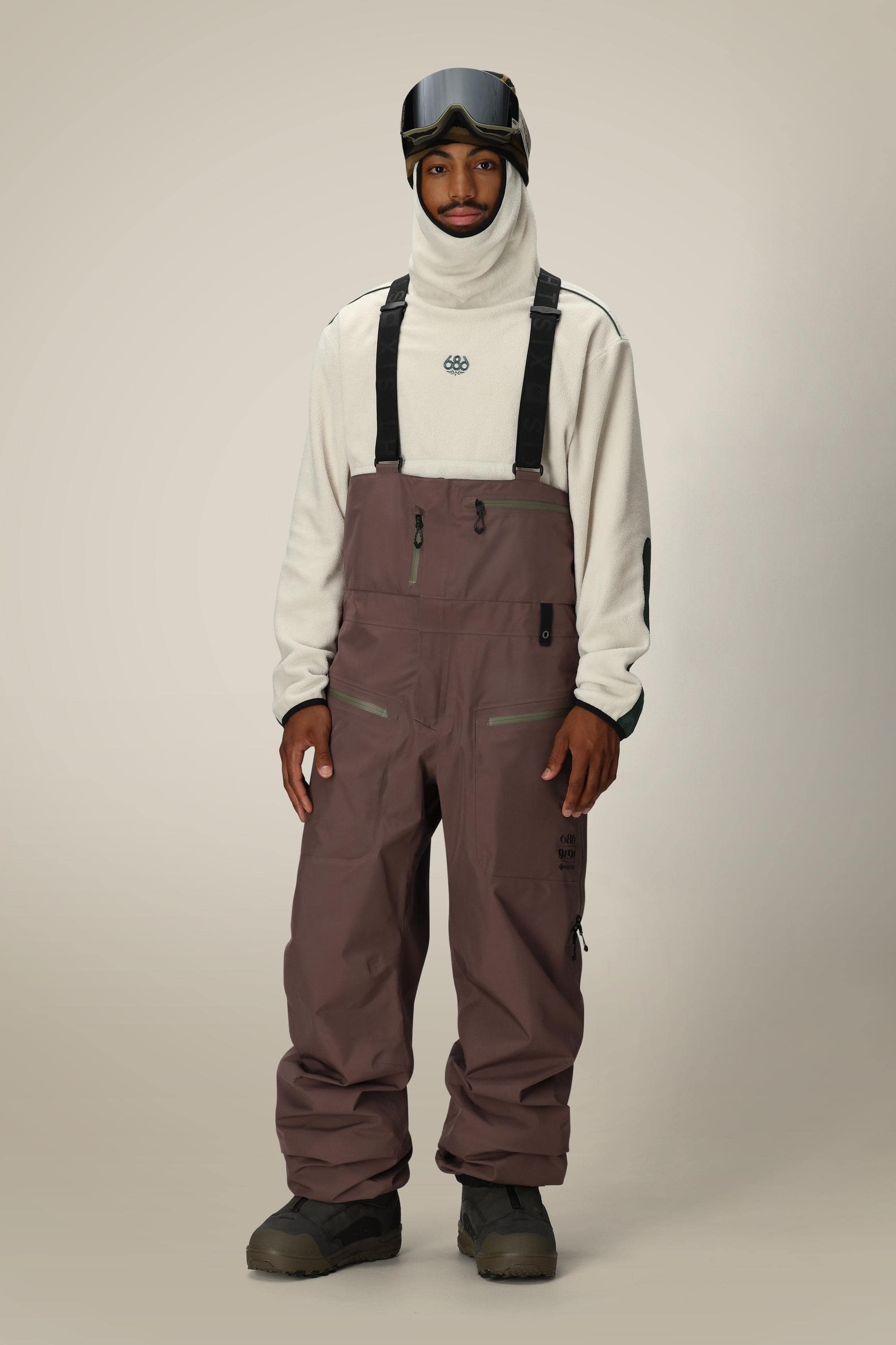 A person stands facing forward against a plain beige background, wearing the 686 Mens GORE-TEX 3L ATV Bib in mauve with a beige hooded sweatshirt, black gloves, dark snow boots, and ski goggles on their head.