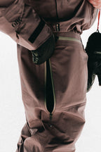 A close-up of someone wearing the 686 Mens GORE-TEX 3L ATV Bib in brown, featuring a large side zipper partially open to show the inner lining, paired with black gloves.