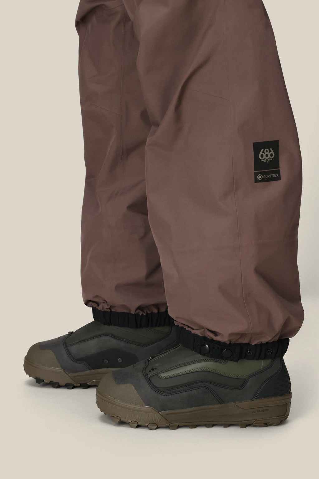 A person is shown from the knees down wearing the 686 Mens GORE-TEX 3L ATV Bib in brown, featuring elastic cuffs, over green and brown outdoor shoes against a neutral background.