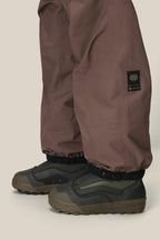 A person is shown from the knees down wearing the 686 Mens GORE-TEX 3L ATV Bib in brown, featuring elastic cuffs, over green and brown outdoor shoes against a neutral background.