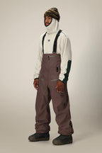 A man stands against a plain light background wearing a beige balaclava, striped beanie, off-white fleece top with dark green accents, and loose-fitting 686 Mens GORE-TEX 3L ATV Bib snow pants with suspenders from 686.