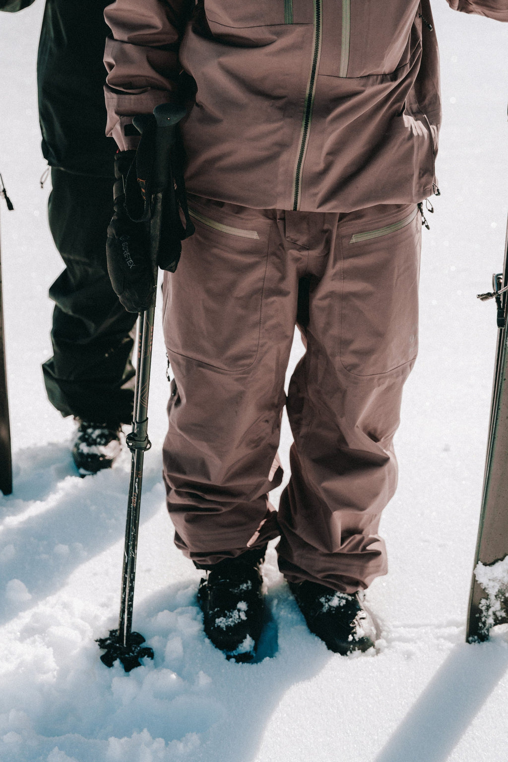 Two people stand in the snow, their lower bodies visible as they wear 686 Mens GORE-TEX 3L ATV Bibs and winter jackets, holding ski poles next to their skis.