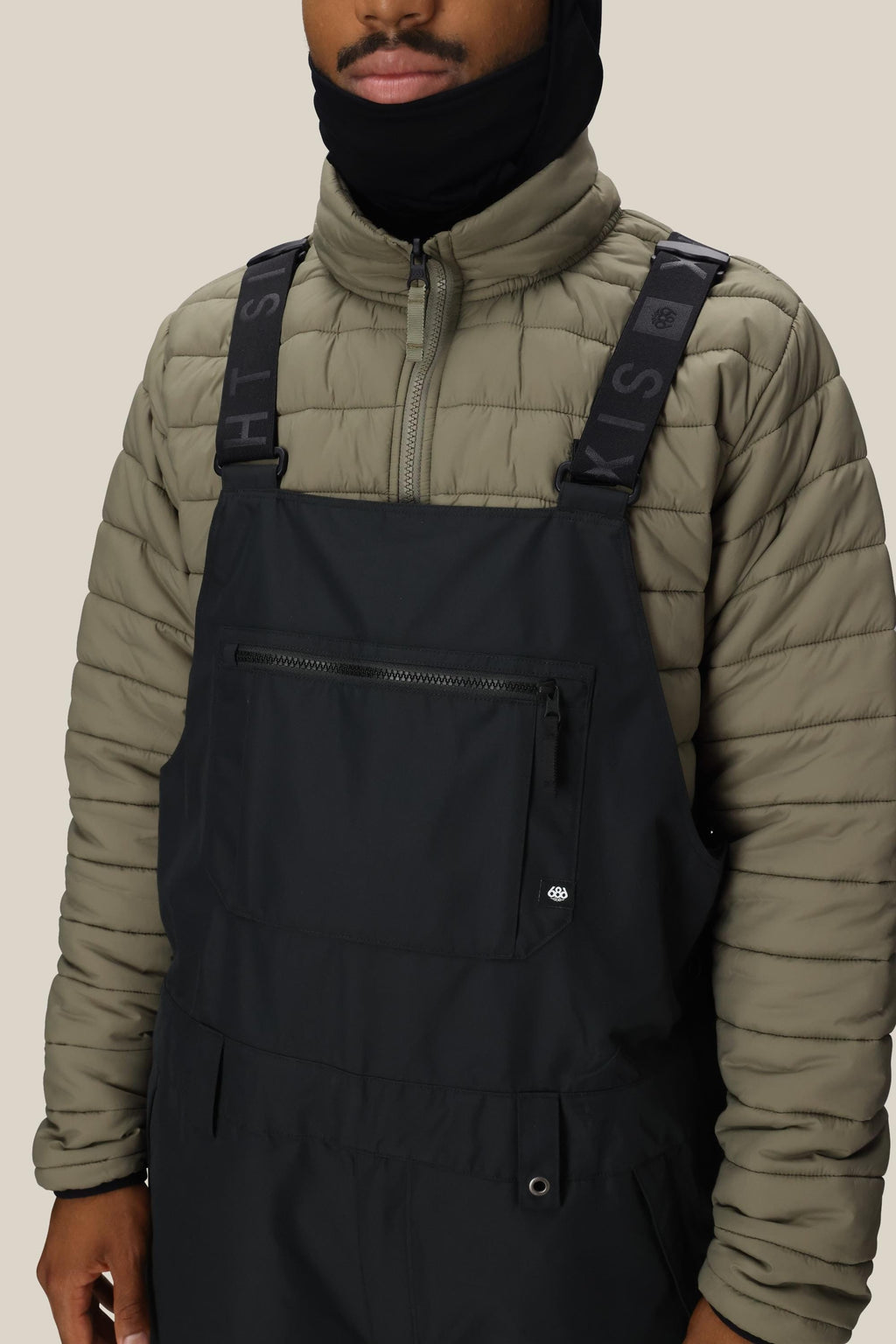 A person wears a light olive-green puffer under the 686 Men's 20K Hot Lap™ Shell Bib by 686, featuring black polyester fabric, zippered chest pocket, and wide straps, with a black face covering against a plain beige background.