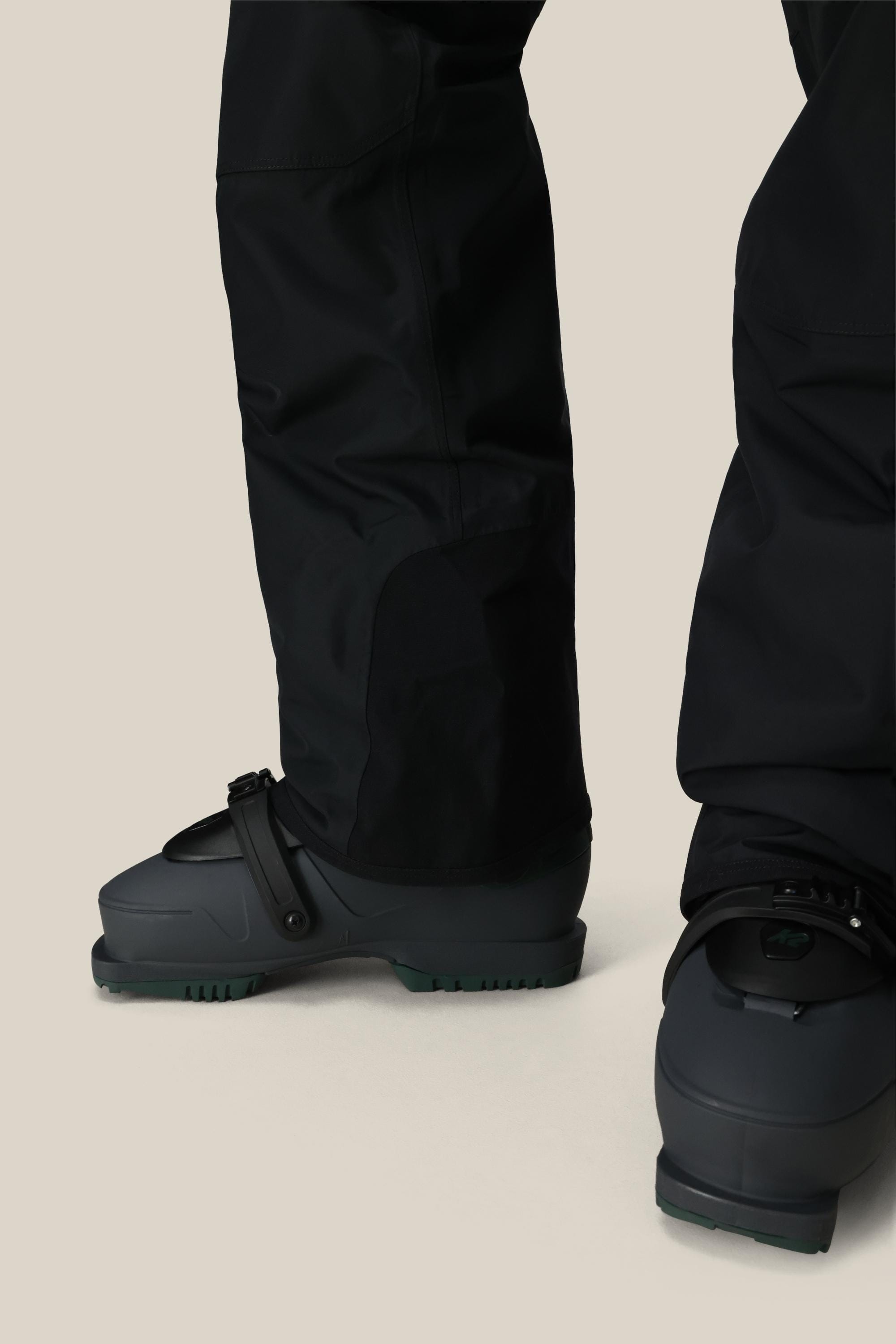 Close-up of a person wearing 686 Men's 20K Hot Lap™ Shell Bib snow pants and dark snowboarding boots with bindings, standing on a light beige background.