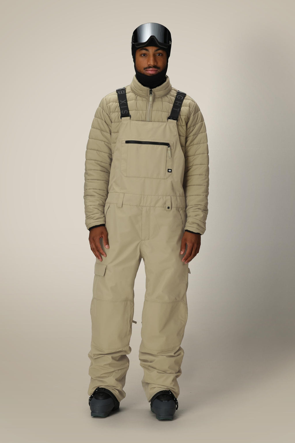 A person wears the 686 Men's 20K Hot Lap™ Shell Bib by 686 with a matching quilted jacket, black gloves, black ski boots, and a black ski helmet with goggles, standing against a plain light background.
