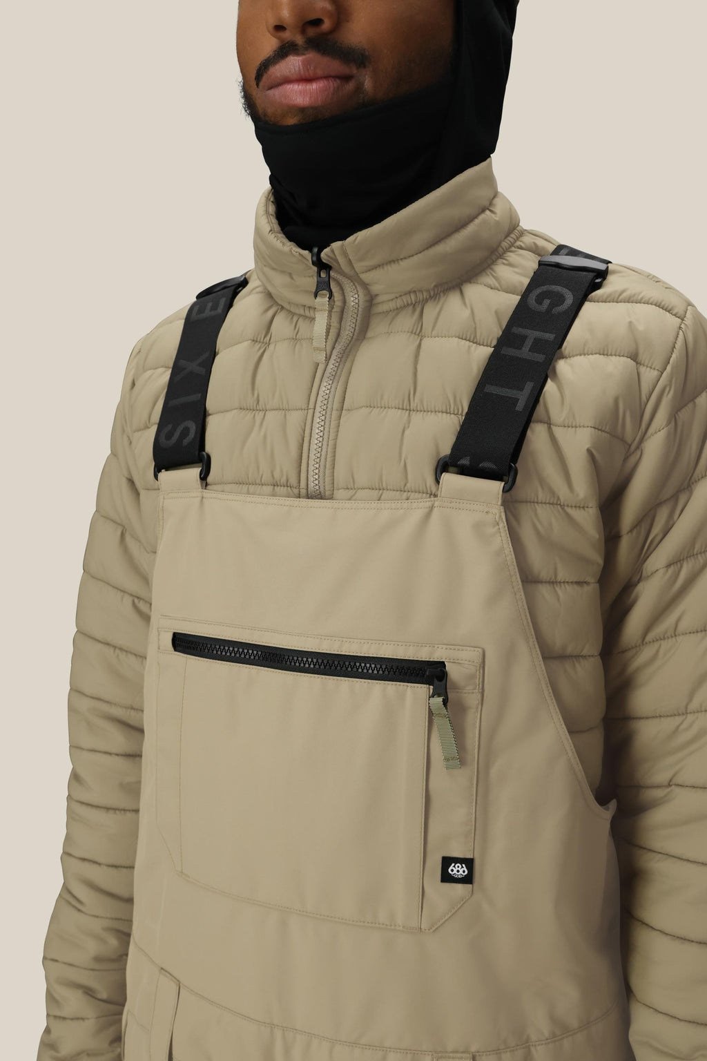 A person wears a beige quilted jacket and 686 Men's 20K Hot Lap™ Shell Bib by 686, featuring black straps and a front zip pocket, partially concealing their face with a black hood or balaclava against a plain background.