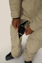 A person in beige 686 Men's 20K Hot Lap™ Shell Bib pants and black snow boots unzips an inner thigh vent, revealing the black mesh lining.