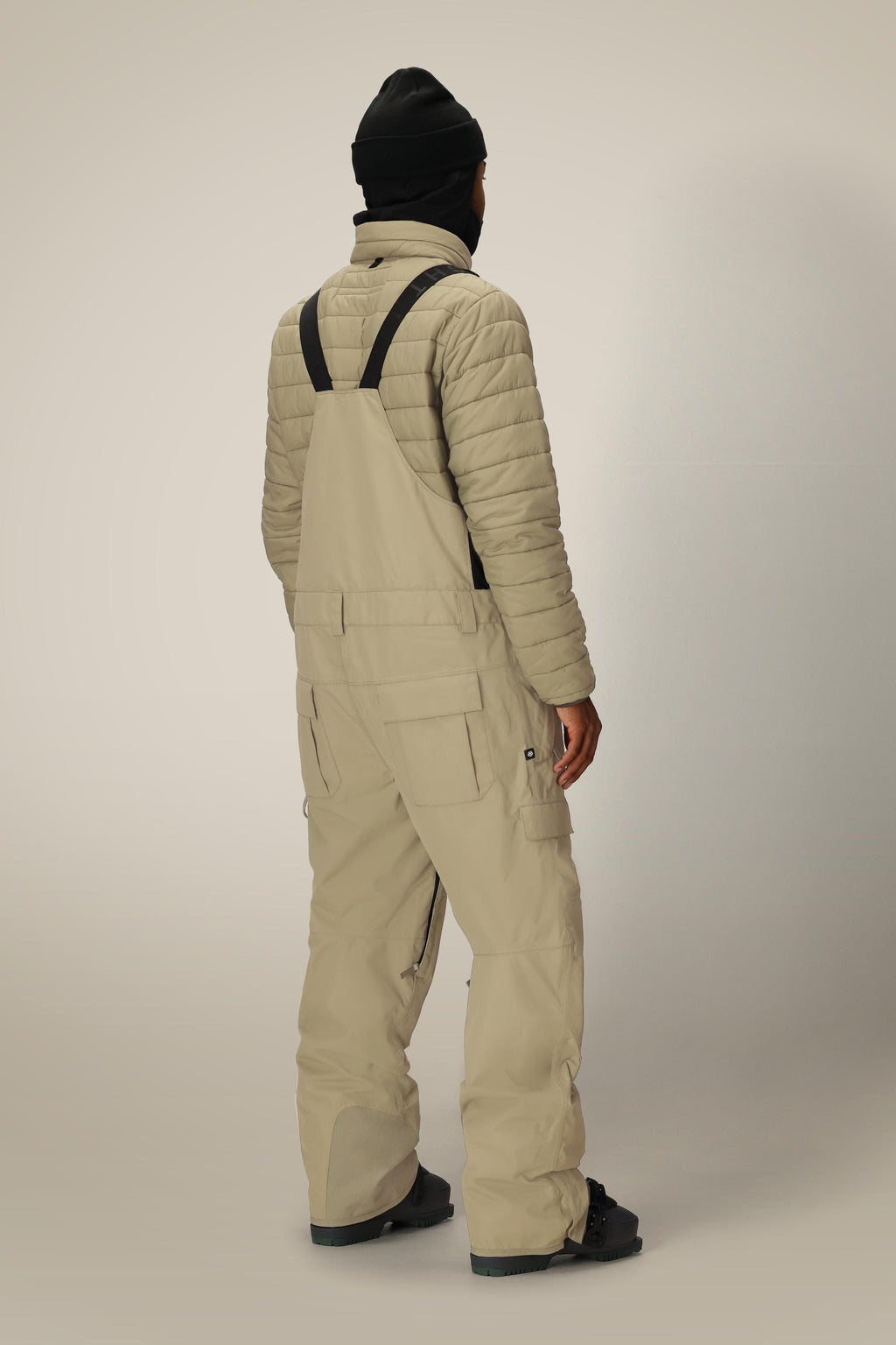 A person wearing a black beanie, a light quilted jacket, and 686 Men's 20K Hot Lap™ Shell Bib from 686 with black straps stands facing away from the camera against a neutral background.