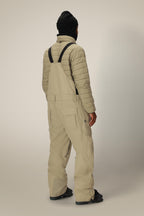 A person wearing a black beanie, a light quilted jacket, and 686 Men's 20K Hot Lap™ Shell Bib from 686 with black straps stands facing away from the camera against a neutral background.