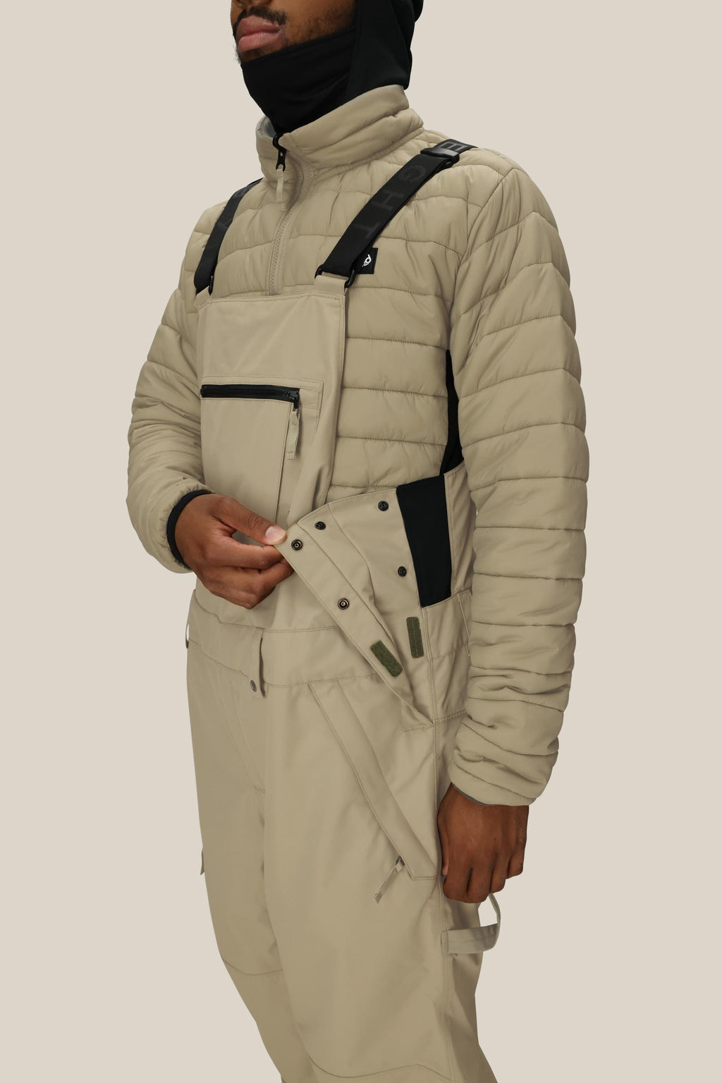 A person, face partly covered by a black hood, fastens the side buttons of beige 686 Men's 20K Hot Lap™ Shell Bib overalls with black straps, paired with a beige quilted jacket.