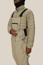 A person, face partly covered by a black hood, fastens the side buttons of beige 686 Men's 20K Hot Lap™ Shell Bib overalls with black straps, paired with a beige quilted jacket.