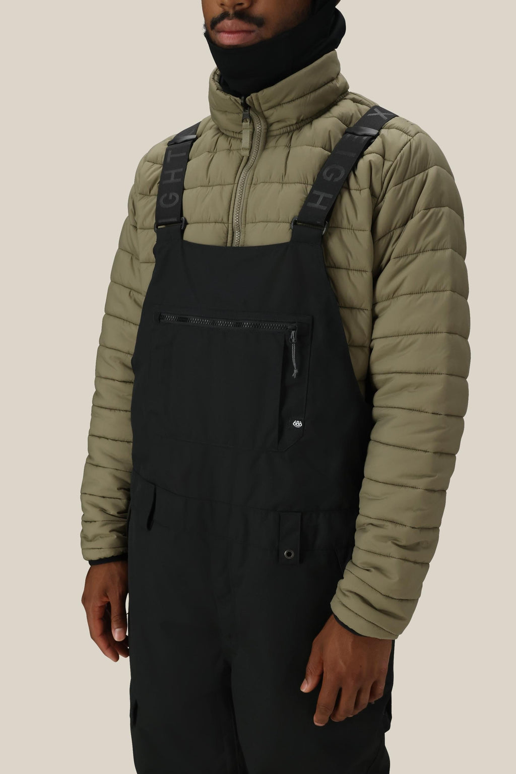 A person stands against a beige background wearing the 686 Men's Hot Lap™ Insulated Bib over a light olive green puffer jacket, with adjustable straps. Their face is partially covered by a black neck gaiter.