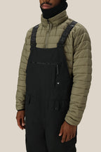 A person stands against a beige background wearing the 686 Men's Hot Lap™ Insulated Bib over a light olive green puffer jacket, with adjustable straps. Their face is partially covered by a black neck gaiter.