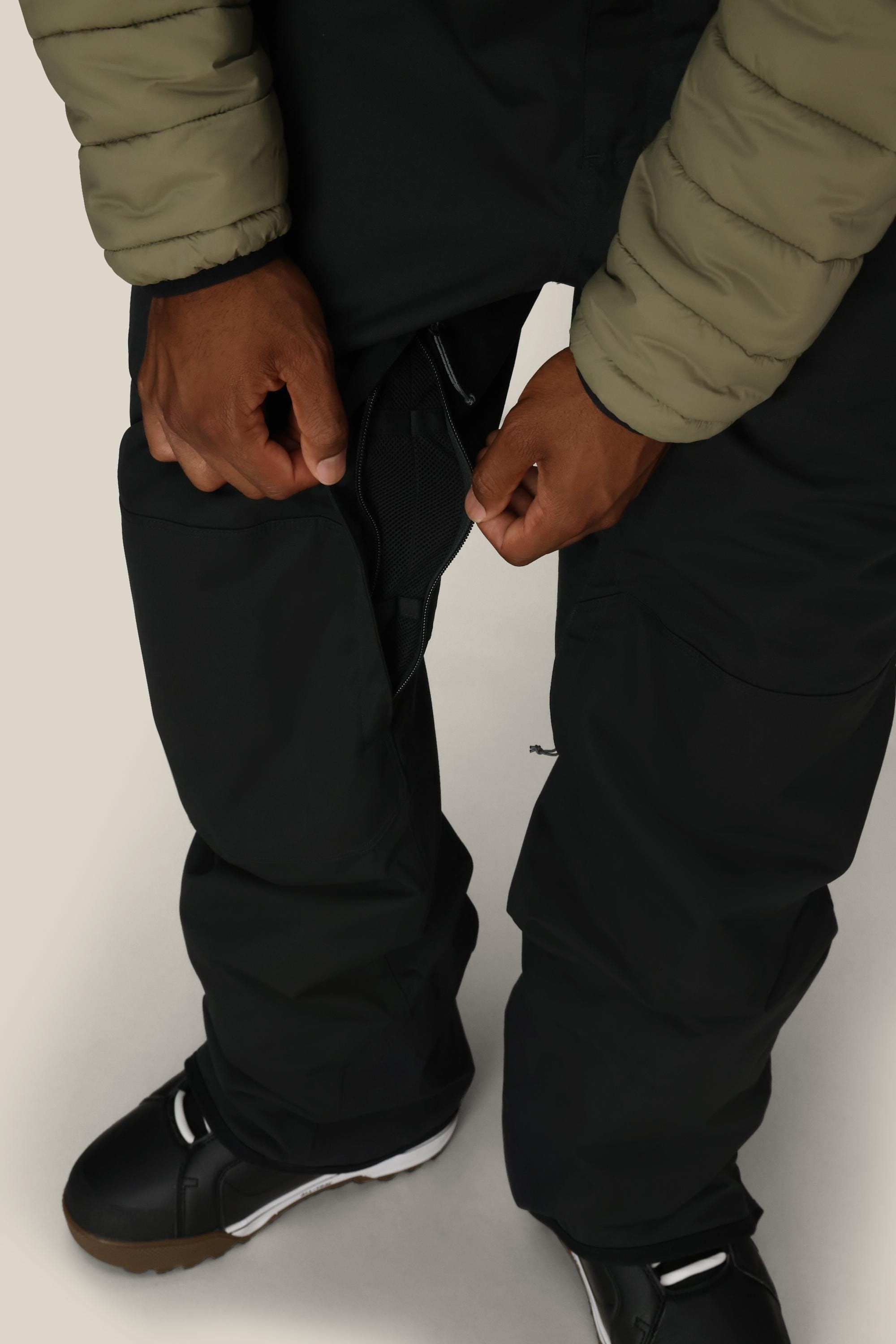 A person in the 686 Men's Hot Lap™ Insulated Bib by 686, made from polyester nylon spandex, unzips a thigh ventilation zipper while wearing black pants and black-and-white shoes.