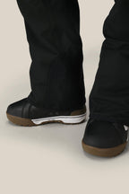 Close-up of a person wearing the 686 Men's Hot Lap™ Insulated Bib by 686—black, waterproof woven snow pants—paired with black snow boots with brown soles, standing on a light-colored surface.