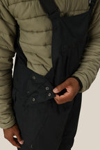 A person wearing the 686 Men's Hot Lap™ Insulated Bib by 686 fastens metal snaps on the side of the black polyester-nylon-spandex overalls with one hand, paired with a green quilted jacket.