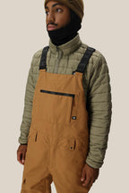 A person wearing the 686 Men's Hot Lap™ Insulated Bib by 686, an olive green puffer jacket, a tan beanie, and a black neck gaiter stands against a plain light background.