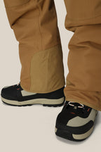 A close-up of someone in tan pants and black-and-beige outdoor shoes stands on a light background—perfectly paired with the 686 Men's Hot Lap™ Insulated Bib by 686, crafted from waterproof woven polyester, nylon, and spandex.
