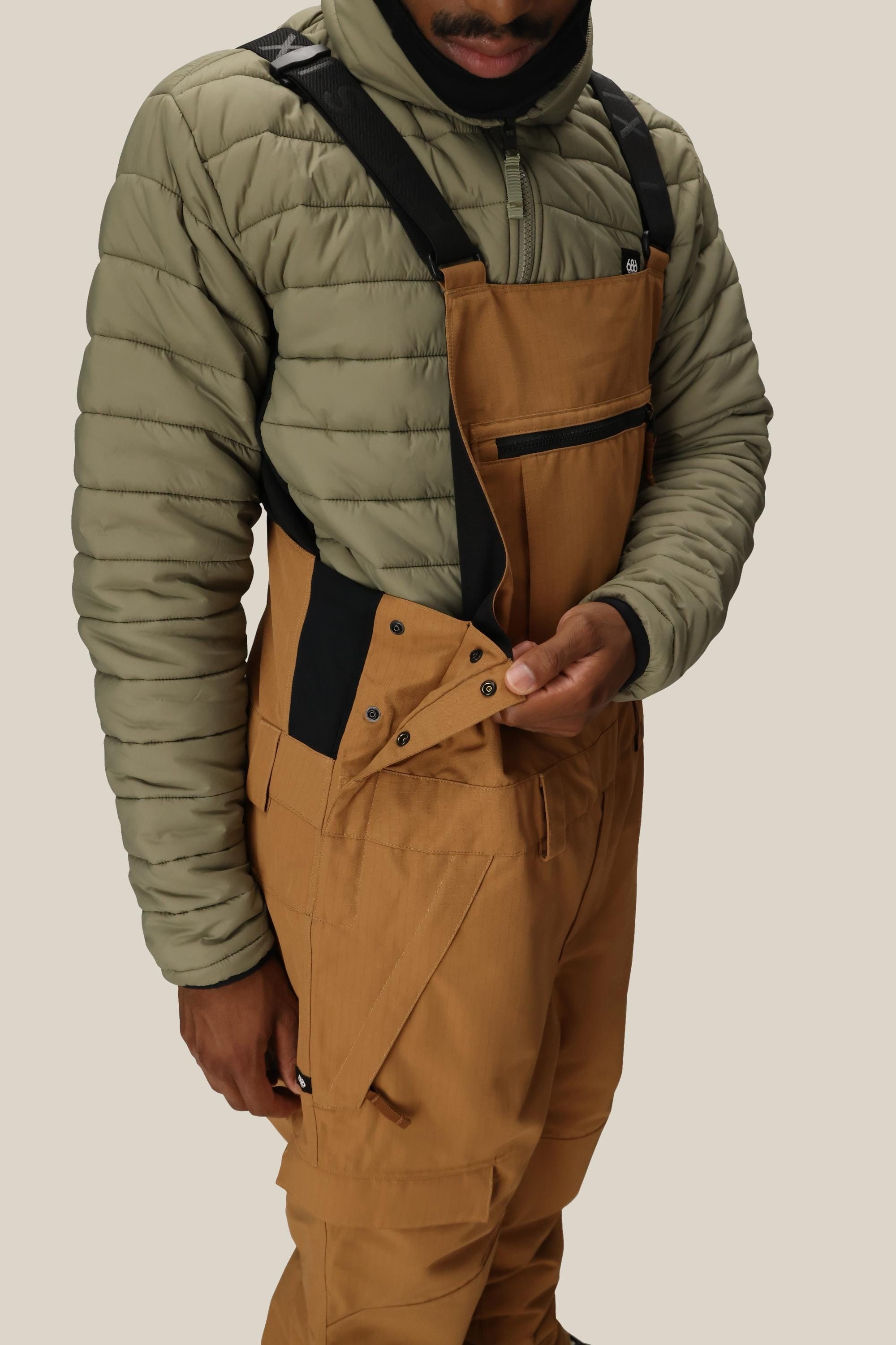 A person wearing a light green quilted jacket and 686 Men's Hot Lap™ Insulated Bib by 686, unsnapping the side buttons, with a plain neutral background.