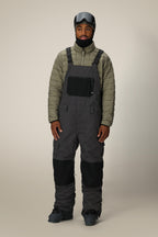 A man stands against a plain beige background wearing the 686 Men's Hot Lap™ Insulated Bib, a gray insulated bib made from polyester nylon spandex with black knee patches, paired with black boots, gloves, and a black ski helmet with goggles.
